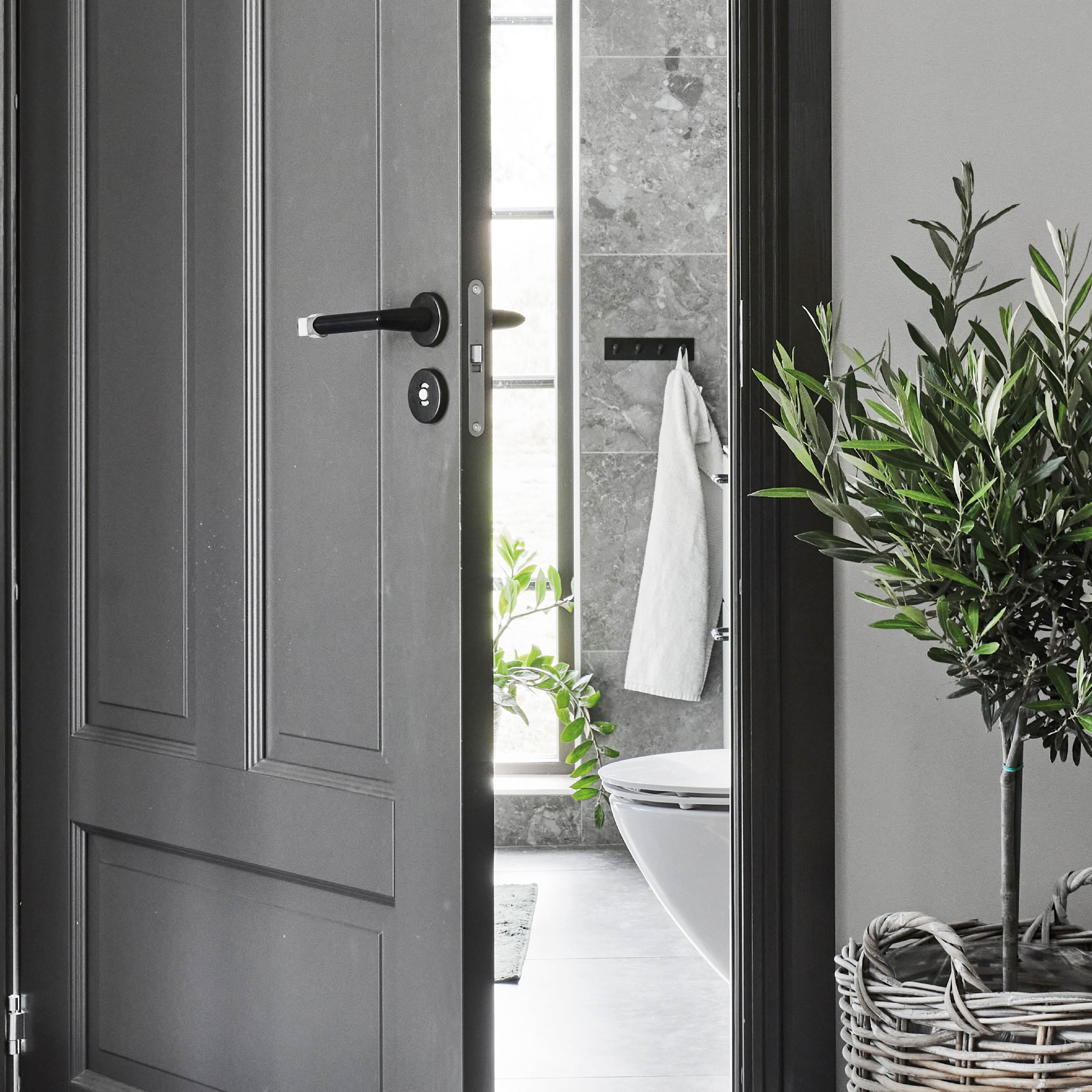 Öppen mörkgrå dörr till modernt badrum med grå kakel, vit handduk, toalett och grön växt i korg.