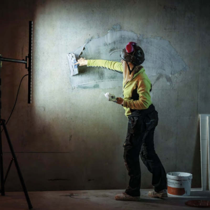 En person i gul tröja och skyddshjälm spacklar en betongvägg framför arbetslampa