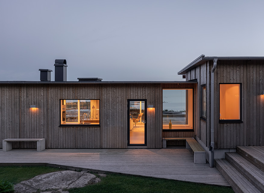 Modernt trähus på träaltan, varmt sken inifrån, havsutsikt genom fönstren i skymning