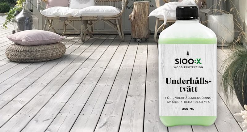 Grön SiOO:X-flaska på grå träterrass med rottingmöbler, rosa dyna, vita kuddar och växter.