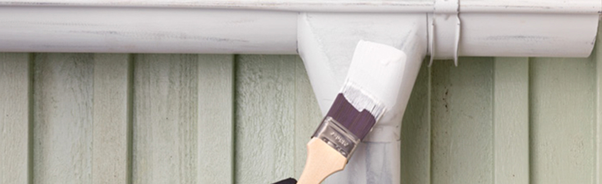 En pensel målar ett vitt stuprör mot ljusgrön vertikal träpanel.