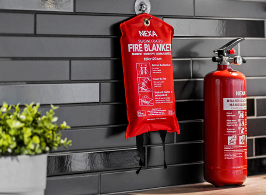 Röd brandskyddsfilt i NEXA-fodral på svart kakelvägg bredvid röd brandsläckare och växt