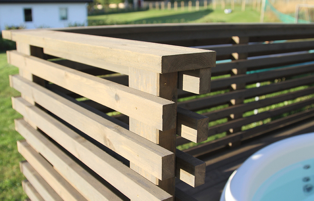 Brun träskärm med horisontella ribbor runt ett vitt spabad i solig gräsmiljö.