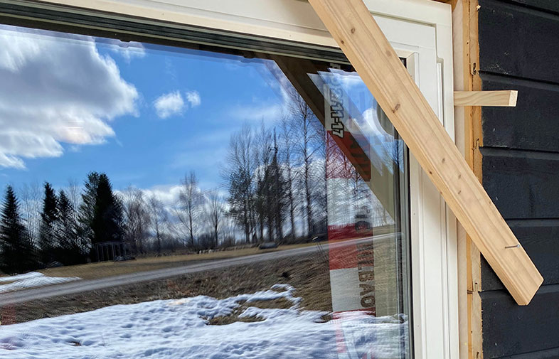 Fönster på husbygge speglar blå himmel, vita moln, nakna träd och snö på marken