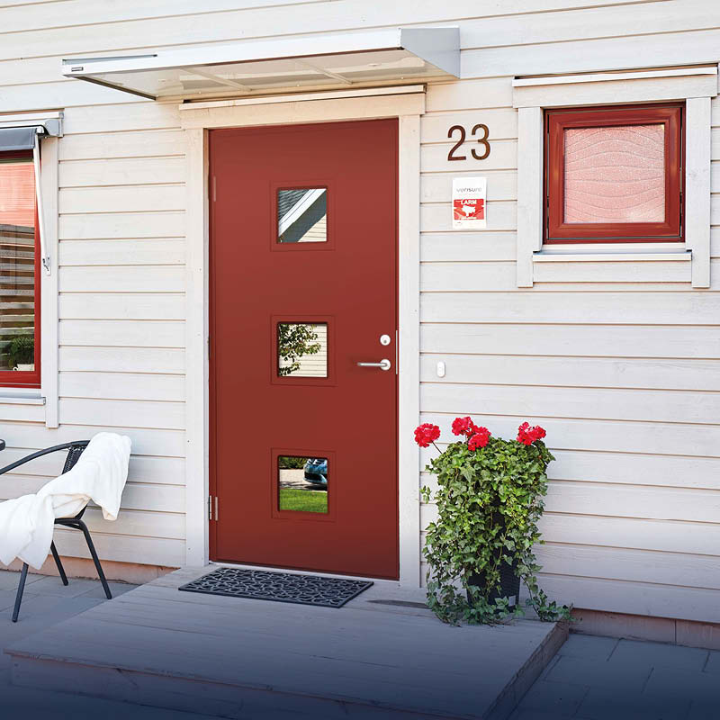 Entré till ljus trävilla med röd ytterdörr, husnr 23, svart stol och röda pelargoner.