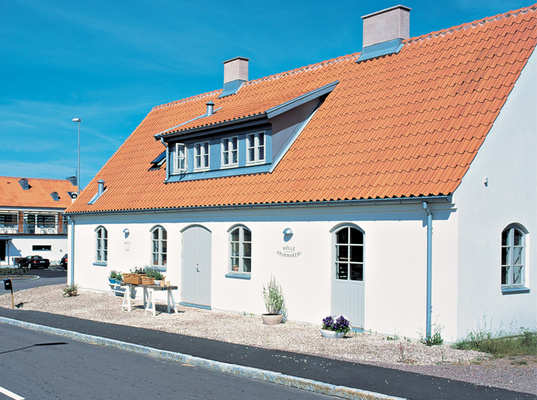 Vit skånelänga med rött tegeltak, loftfönster, utebord och blommor mot klar blå himmel.