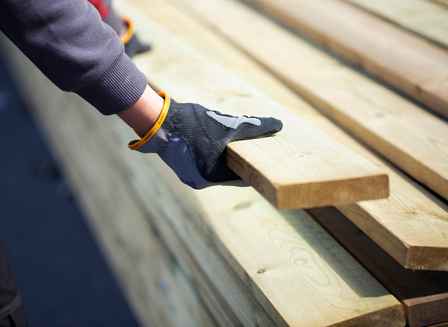 Svartgrå handske med gul kant lyfter en ljus träplanka från stapel av träbrädor.