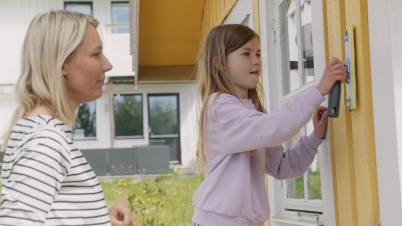 Flicka i lila tröja låser upp gul ytterdörr medan kvinna i randig tröja står bakom.
