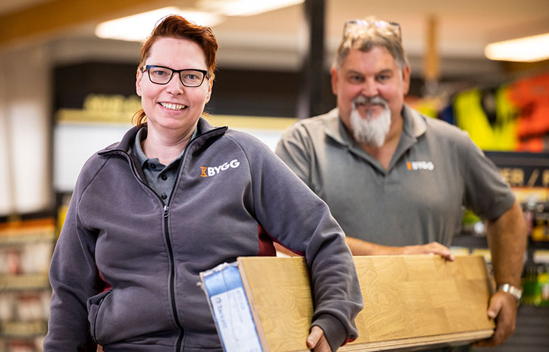 Två anställda i gråa BYGG-tröjor bär plankor i en ljus bygghandel.