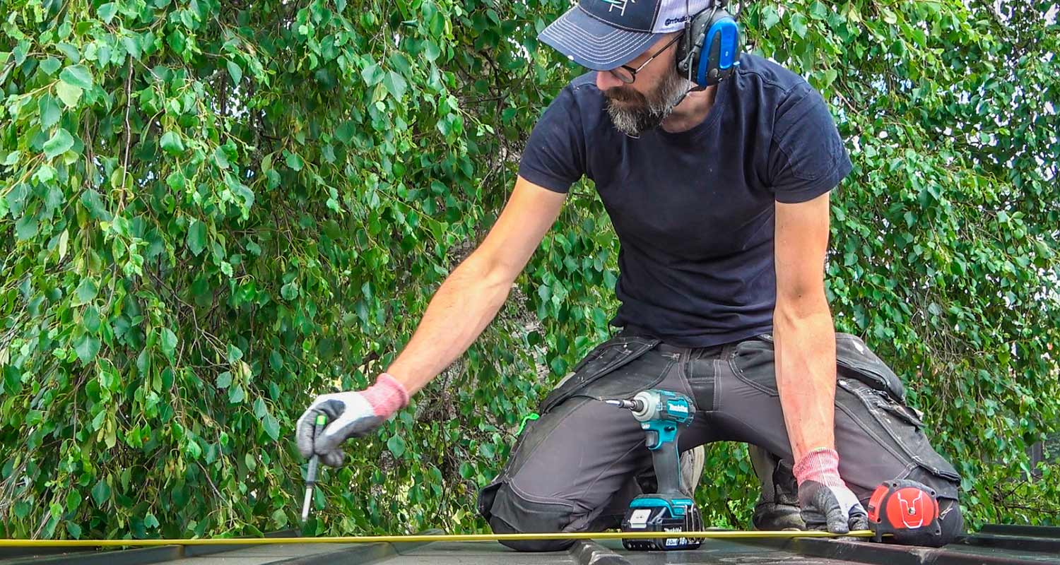 Man i svart tröja med hörselskydd mäter och borrar på ett plåttak framför grön lövverk.
