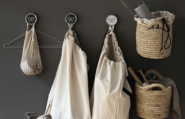 Mörkgrå vägg med tre vita krokar märkta 30°/40°, hänger nät- och tygpåsar samt flätade korgar.