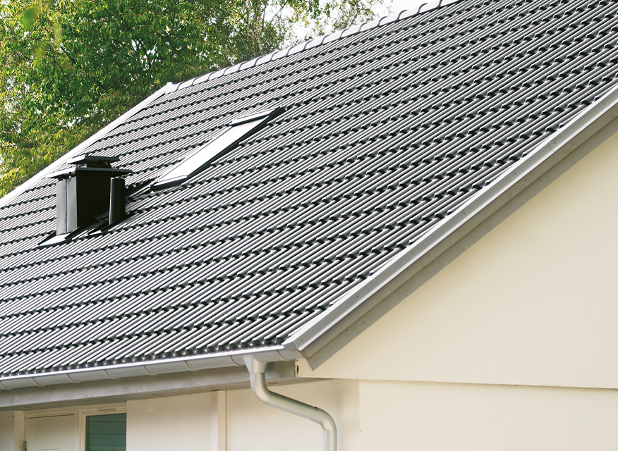 Ett hus med ett lutande tak av profilerad plåt. På taket finns ett takfönster och en svart skorsten/ventilationshuv. Fasaden är ljus och i förgrunden syns en hängränna. Bakgrunden består av träd med gröna blad.