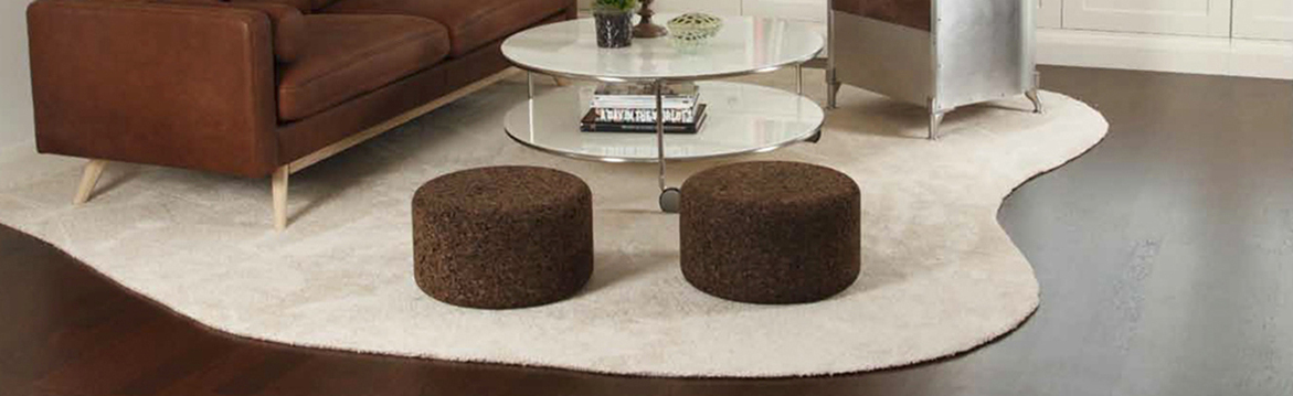 Interiör: brun soffa vid vit oregelbunden matta med två mörkbruna puffar och runt glasbord.