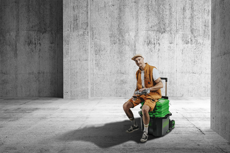 Person i orange arbetskläder sitter på grön-svart verktygslåda i tom grå betonghall.