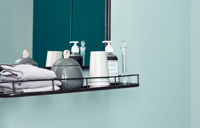 Svart hylla med grå handdukar, keramikkrukor, vit pumpflaska och klar glaskaraff mot turkos vägg