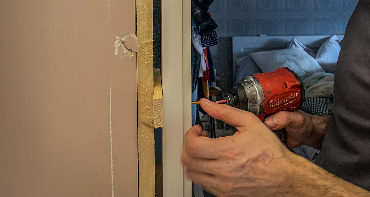 Händer använder röd skruvdragare på vit dörrkarm mot beige vägg, sovrum i bakgrunden.