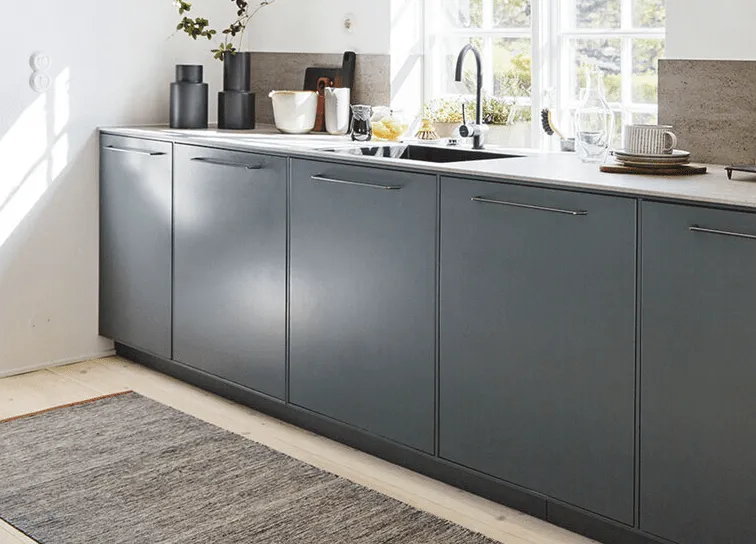 Solbelyst modernt kök med mörkgrå skåpluckor, ljus bänkskiva, diskho och redskap.