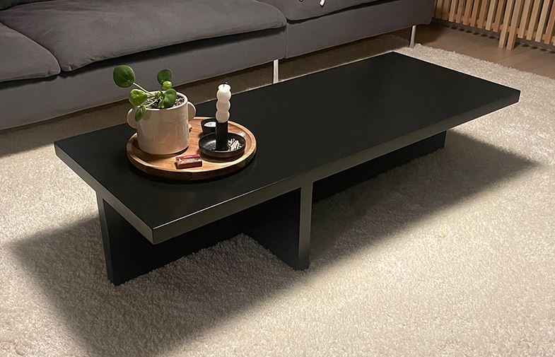 Svart minimalistiskt soffbord på vit matta med träbricka, grön krukväxt och vit spiralljus.