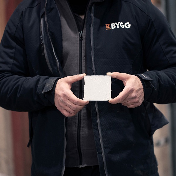 Person i svart K Bygg-jacka håller vit kakelplatta med båda händerna