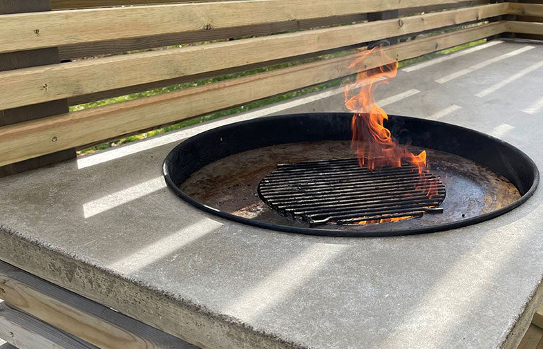 Lågor i rund svart grillgrop på grå betongyta framför trästaket.