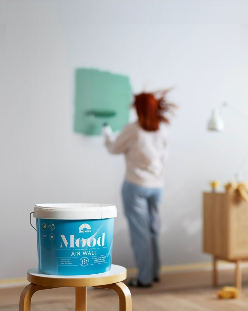 Blå Beckers Mood Air Wall-burk på träpall, suddig kvinna målar vit vägg mintgrön.