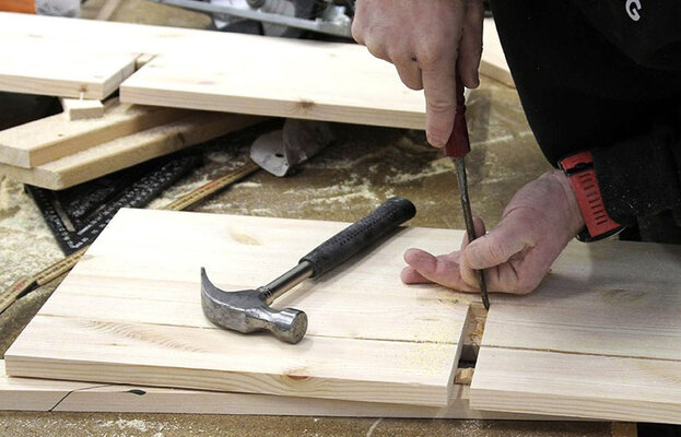 Händer mejslar ljust trä på dammig arbetsbänk med hammare, rött handtag och sågspån.
