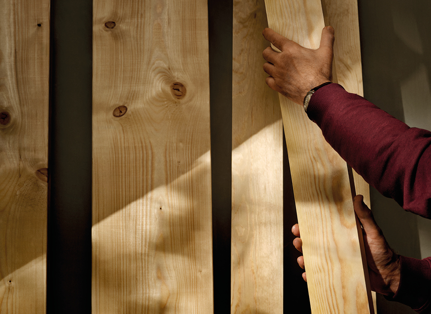 Hand i vinröd tröja håller ljus träplanka framför solbelysta vertikala träpaneler