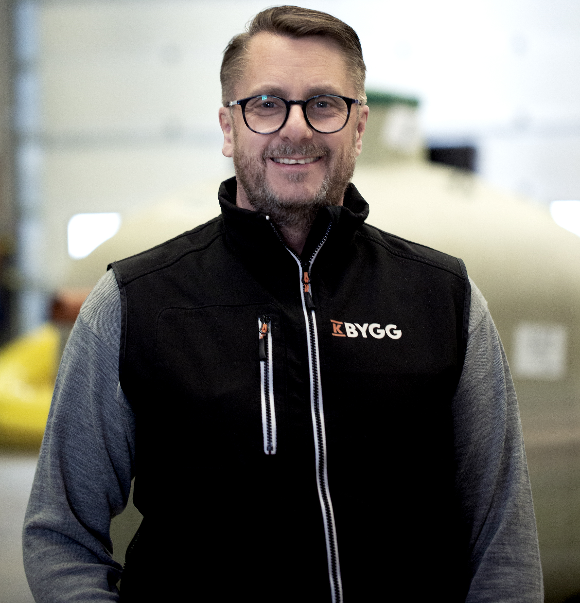 Man i svart byggväst med KBYGG-logotyp, grå tröja i verkstadsmiljö med stor tank bakom