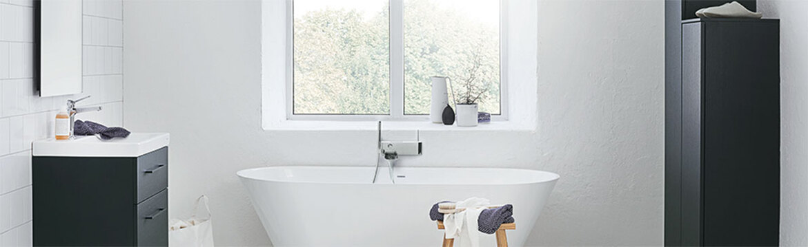 Modernt badrum med fristående vit oval badkar framför fönster, svart skåp, träpall med handdukar.