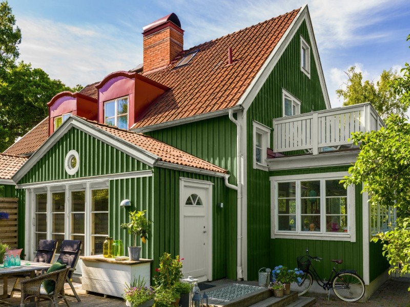 Grönt hus med rött tegeltak, vita fönster, balkong, uteplats med bord, stolar och växter