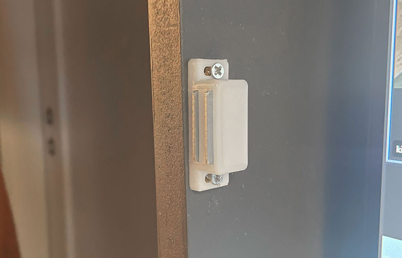 Vit plastdörrsensor med skruvar monterad på mörkgrå dörrstomme intill träfoder.