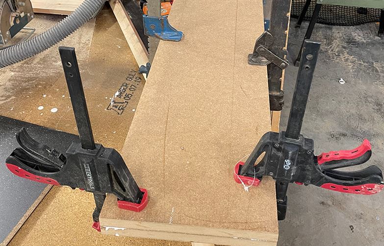 Svarta och röda snabbklämmor håller en spånskiva på plats i verkstad med såg i bakgrunden.