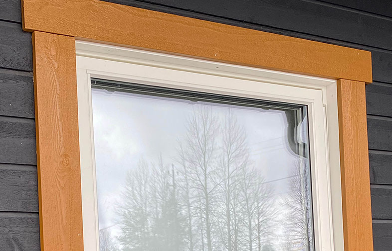 Vitt fönster med orangebruna lister i svart träfasad, glas speglar nakna träd.