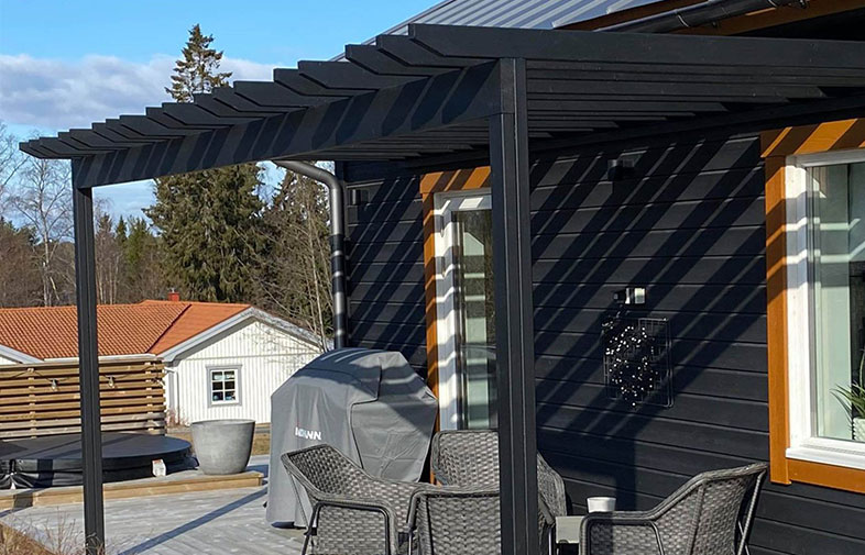Svart pergola på träaltan med randiga skuggor, grå gasolgrill och utemöbler