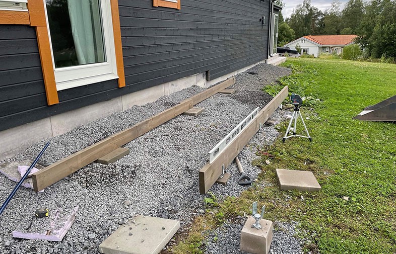 Underlag med grus, träbalkar och lasernivå vid svart fasad med orange fönsterfoder.