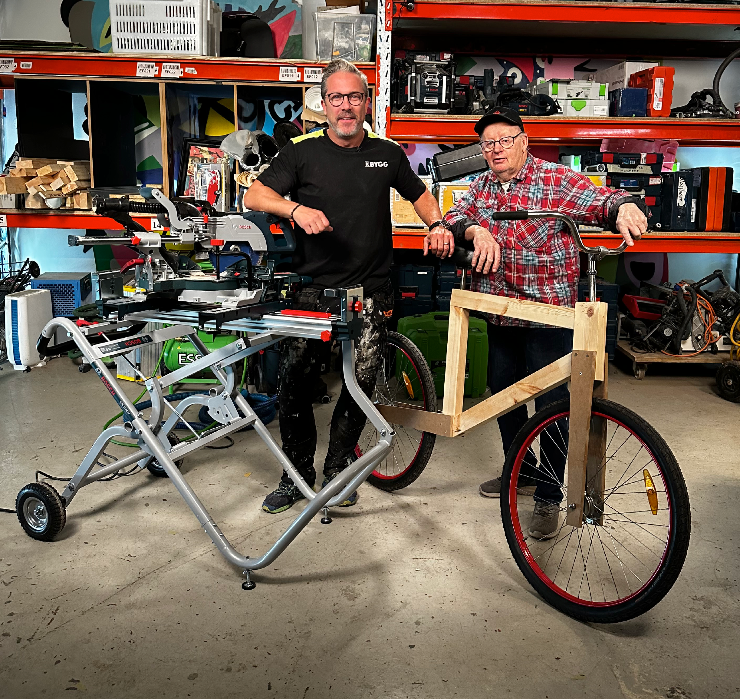 Två hantverkare i verkstad med grå bänksåg och hembyggd träcykelram med röda hjul, verktygshyllor