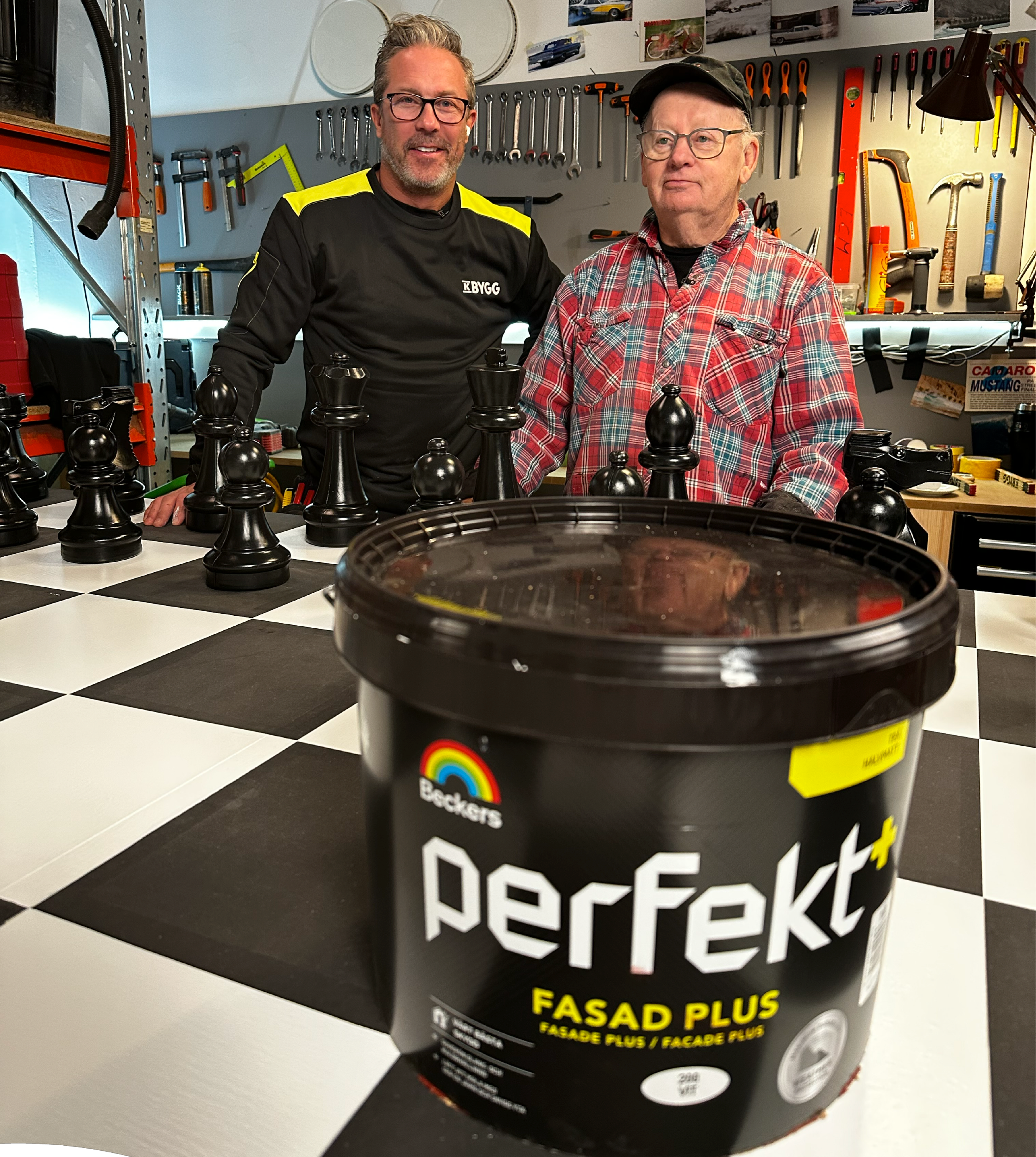 Två män vid stor svartvit schackbräda i verkstad, i förgrunden svart Beckers Perfekt-färgburk.