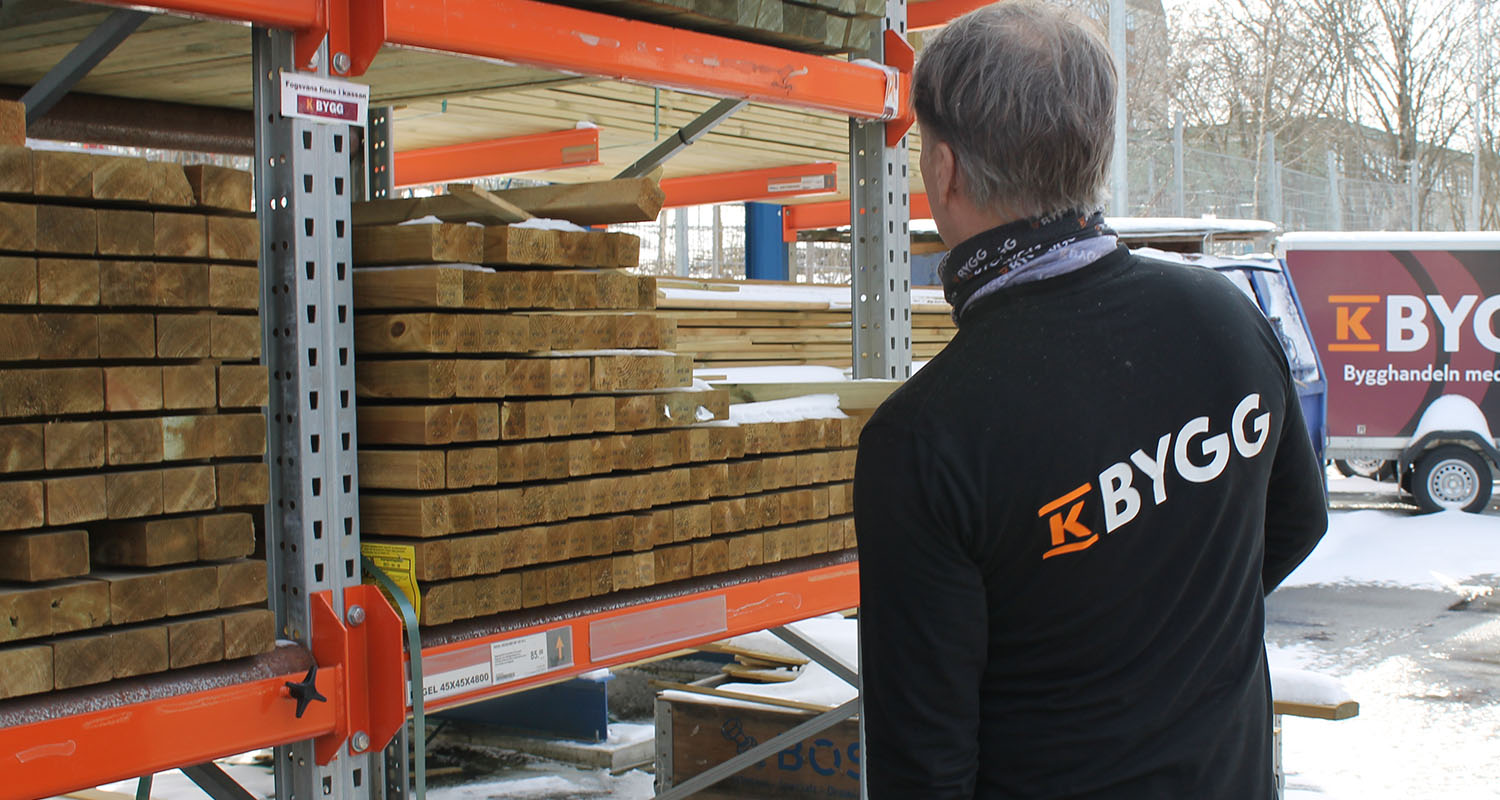 Man i svart K-BYGG-tröja granskar staplade träbalkar på orange hyllor utomhus i snö