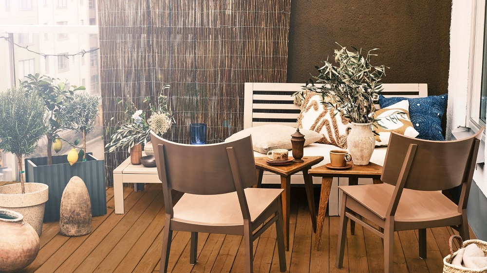 Solig balkong med trämöbler (vit bänk, bruna stolar), färgglada kuddar och gröna växter