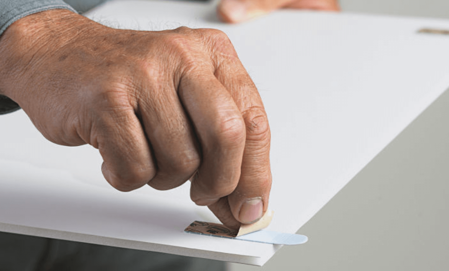 Närbild på hand som drar bort skyddsremsa från blå dubbelhäftande tejp på vit skiva.