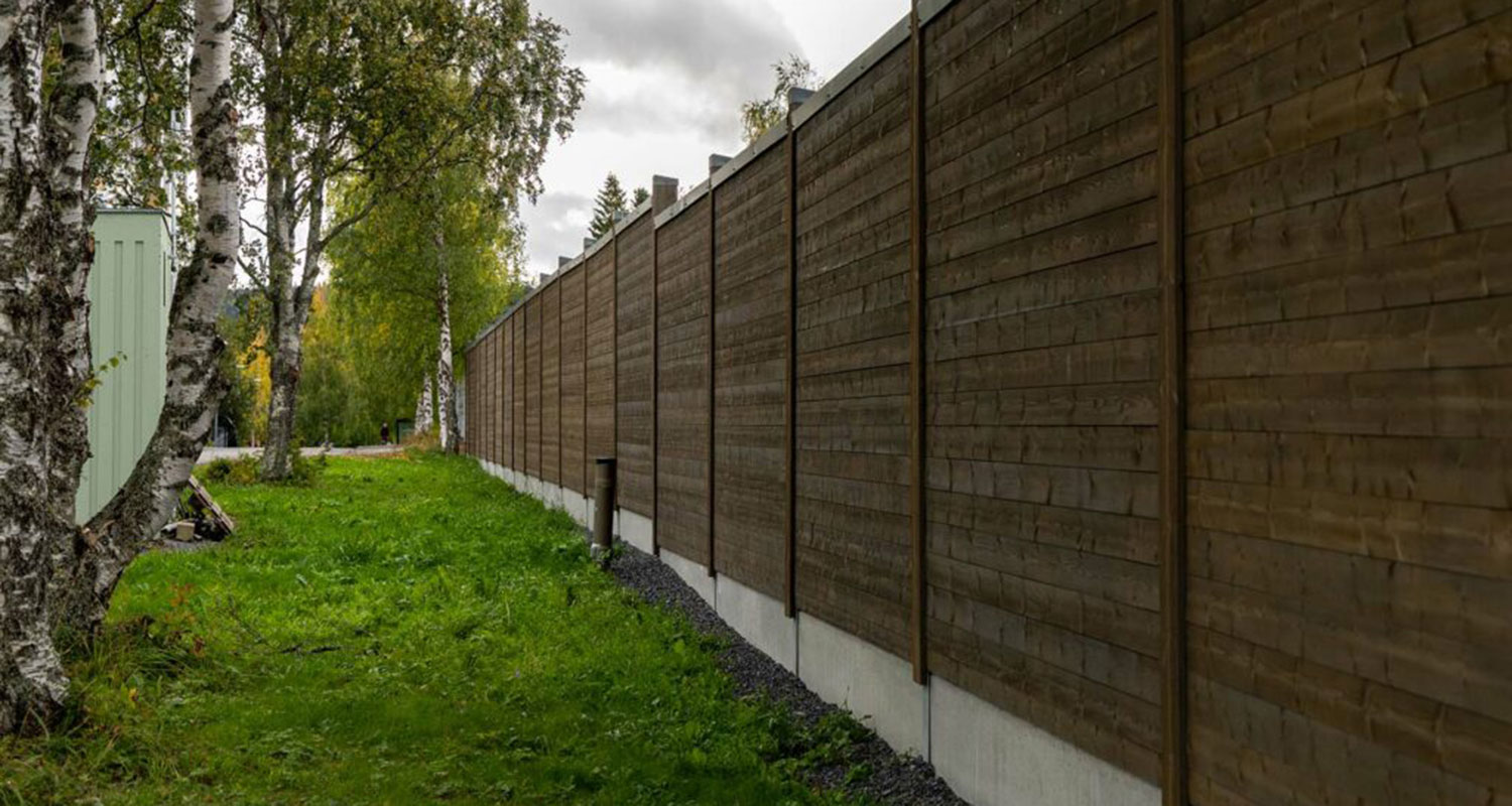 Långt brunt trästaket längs grön gräsyta med björkar under molnig himmel