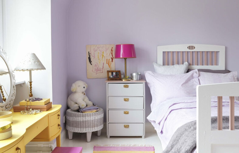 Barnrum i pastell: lila vägg, vit säng med ljuslila sängkläder, rosa lampa, gult skrivbord.