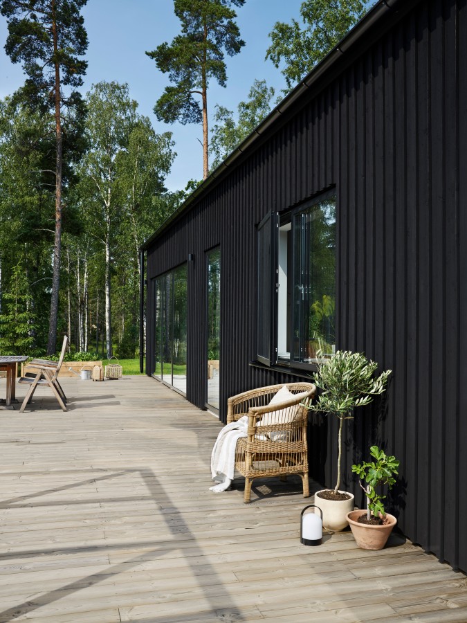 Svart träfasad med stora glasdörrar, ljust trädäck, rottingstol och krukväxter mot skog