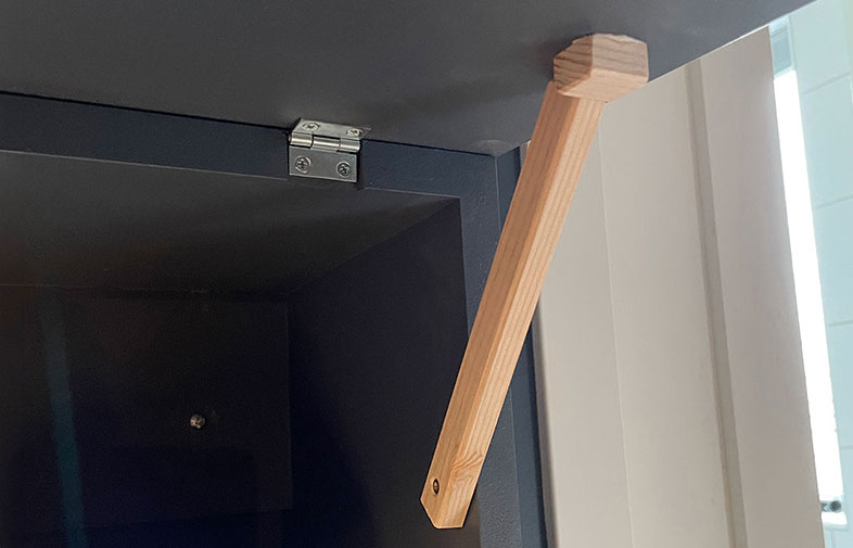 Mörkgrått skåp med öppen lucka som stöds av en ljusträkil, vitt fönster i bakgrunden.