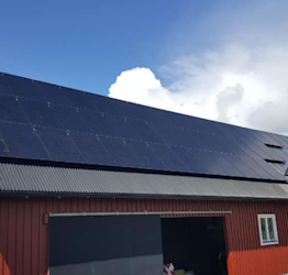 Röd ladugård med mörkblå solpaneler på taket, blå himmel och vita moln.