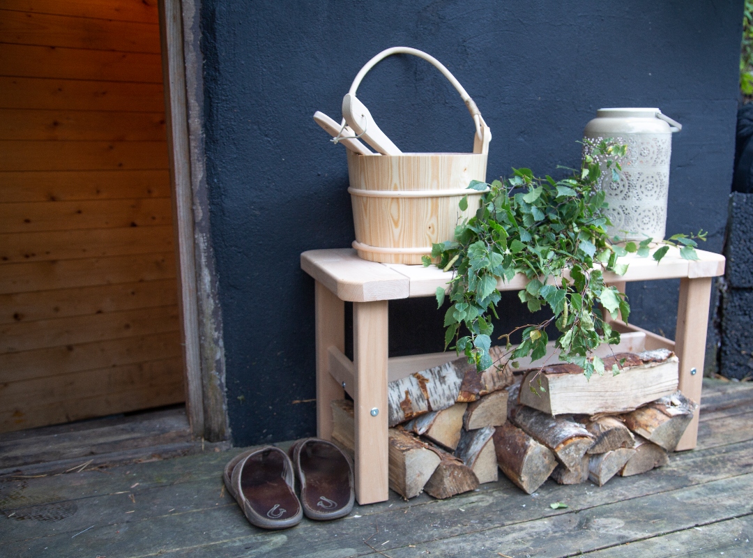 Bastubänk utanför svart vägg med ved, bastuvihta, trähink, keramikkanna och tofflor