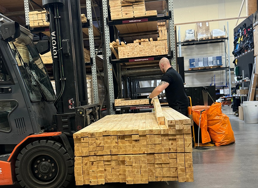 Man i svart tröja lastar träreglar på gaffeltruck framför lagerhyllor, orange säckar vid sidan.