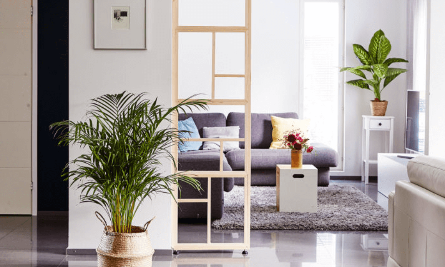 Ljust vardagsrum med grå soffa, färgglada kuddar, gröna växter och bukett på vitt bord.