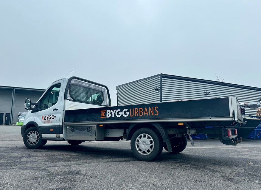 Vit flakbil med svart flak märkt 'KBYGG Urbans' står på asfalt framför industribyggnad under grå himmel