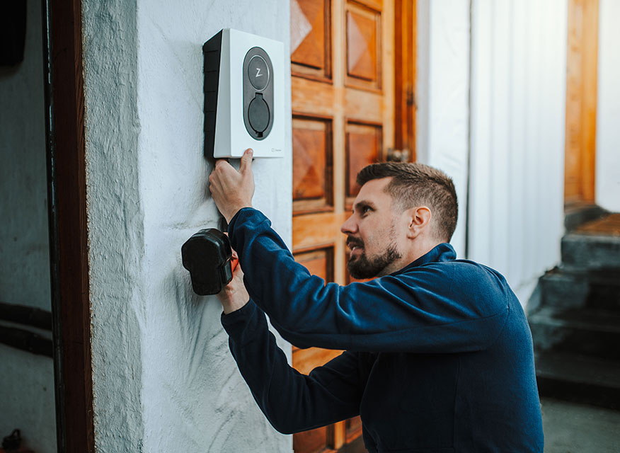 Man i blå tröja monterar svartvit elbilsladdare på vit vägg framför bruna dörrar.